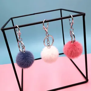 Three colorful plush pom pom keychains in pink, white, and blue on a black stand.