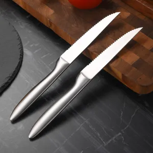 Two stainless steel serrated steak knives on a wooden cutting board and slate surface.