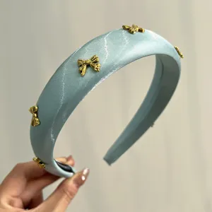 Detail view of a light blue satin padded headband with gold-toned metal bow charms.