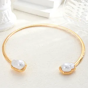 Primer plano de un collar rígido dorado decorado con dos perlas barrocas de imitación.