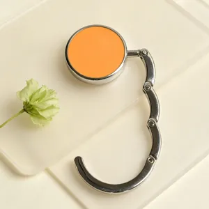 Foldable metal bag hook with orange circular top shown in detail on a minimalist desk