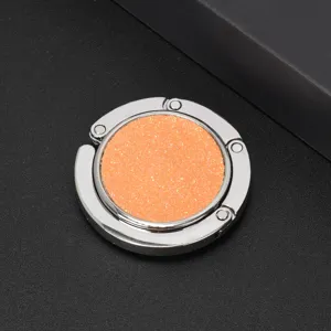 Close-up of circular folding bag hook with orange glitter inlay and silver metal frame.