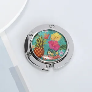 Close-up view of a circular foldable metal handbag hook with a tropical pineapple pattern.