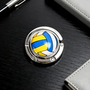 Faltbarer Taschenhaken aus Metall mit buntem Volleyballmuster auf einem dunklen Tisch.