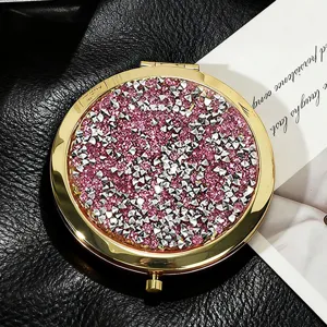 Close-up of a round golden makeup mirror with pink and silver rhinestone lid on black leather.