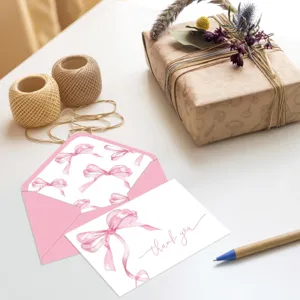 Detalle de tarjeta de agradecimiento con lazo rosa de acuarela y sobre a juego en mesa.