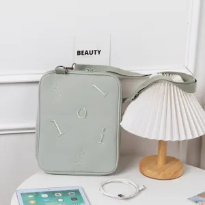 A sage green embroidered iPad messenger bag on a white desk next to a pleated table lamp