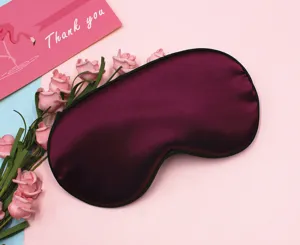 Close-up of a burgundy satin sleep eye mask with black piping on a pink backdrop