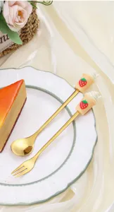 Primer plano de lindo mango de taza de té de leche con fresa en cuchara y tenedor dorados.