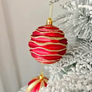 Nahaufnahme einer roten Weihnachtskugel mit goldenen Glitzerwellen an verschneiten Ästen.