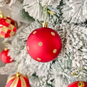 Nahaufnahme einer roten Weihnachtskugel mit goldenen Pünktchen an verschneitem Tannenbaum.
