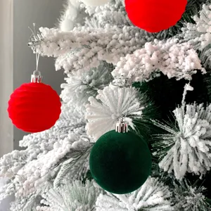 Nahaufnahme von roten und grünen Samt-Christbaumkugeln an schneebedeckten Ästen.