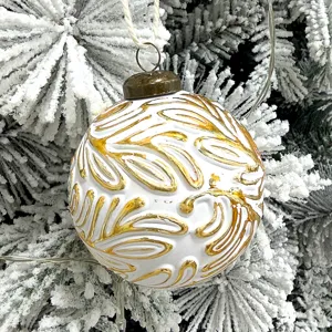 Nahaufnahme einer weiß-gold verziert geprägten Weihnachtskugel am verschneiten Baum.