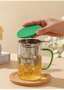 Glass tea infuser cup with stainless steel filter basket, green handle, and wooden coaster.