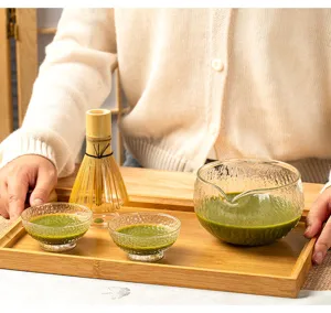 Close-up detail view of clear hammered glass matcha bowl and bamboo whisk on wooden tray