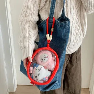 Red plush doll display bag with transparent window hanging on a blue denim tote bag