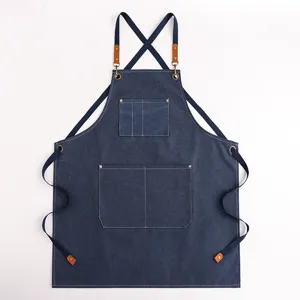 Grembiule in tela di jeans spessa con spalline incrociate, tasche multiple e dettagli in pelle.