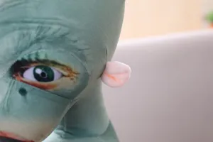 Close-up detail of funny green alien plush toy featuring realistic eye print and cute pink ears.