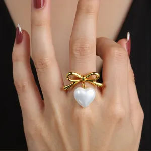Close-up detail of a chic gold bow ring with a dangling white heart pearl charm on hand.