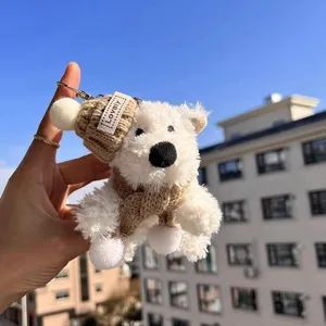 Fluffy white dog plush keychain detail view, featuring a brown knit hat and matching scarf.