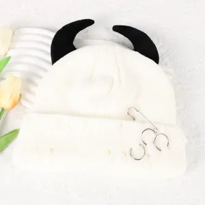 White knit beanie hat with black devil horns, distressed holes, and metal ring pins details.