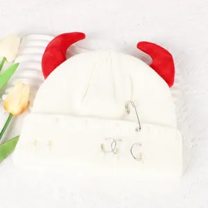 Close up of white knitted beanie with red devil horns and metal safety pins detail.