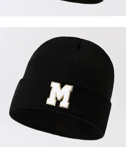 Black knitted beanie hat close-up detail showcasing the white and gold letter M patch design.