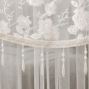 Detalle de primer plano de cortina de encaje blanco con bordado floral y borlas de perlas.