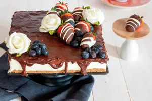Detailed view of square chocolate cake with strawberries, blueberries and white roses on table.
