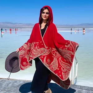 Model wearing a thick red knit hooded shawl with ethnic patterns in a scenic outdoor setting.
