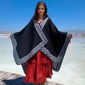 Model wearing a black hooded poncho with Greek key pattern border against blue sky