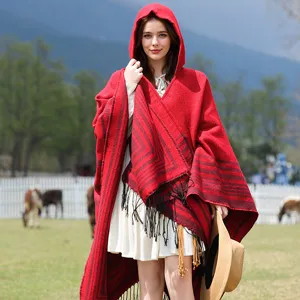 Stylish model wearing red striped hooded knit poncho shawl with tassels in rural field.