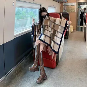 Modischer Damen-Karo-Poncho mit Fransen und braunen Stiefeln in einer Luxus-Zugkabine.