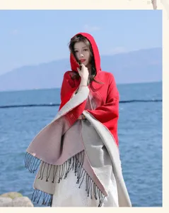 Model draagt een rood-grijze gradiënt sjaal met capuchon en kwastjes bij de blauwe zee