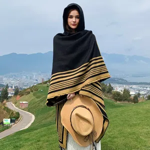 Outdoor view of woman in striped hooded knit poncho shawl holding straw hat on hill