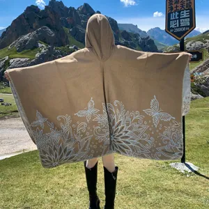 Back view of a hooded khaki knitted poncho with white butterfly floral patterns in a mountain setting