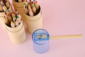 Vista detallada de 12 lápices de colores en tubos kraft con un sacapuntas azul sobre fondo rosa