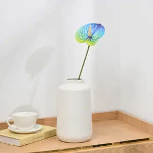Detail view of an artificial blue anthurium flower in a white ceramic vase on wooden desk.