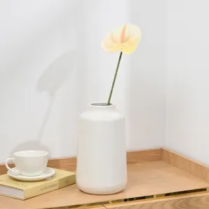 Detailed shot of an artificial anthurium flower in a white ceramic vase on a wooden shelf.