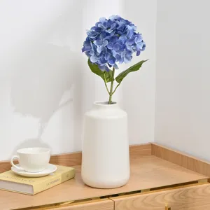 Detailed view of blue artificial hydrangea flower in a white ceramic vase on wooden desk.