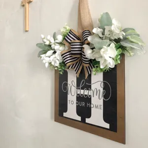 Cartello per porta in legno rustico con lettera H, fiori bianchi e scritta di benvenuto.