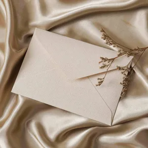 Elegant textured cream envelope on silky champagne fabric background with delicate dry flower.