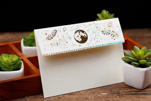 Detailansicht eines eleganten weißen Papierumschlags mit edler Goldfolienprägung und Federmotiv.