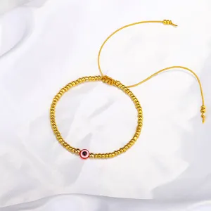 Detailansicht eines goldfarbenen Perlenarmbands mit einem roten bösen Blick auf weißer Seide
