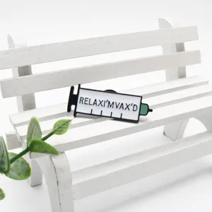 Détail d'une broche en émail en forme de seringue avec le texte 'RELAX I'M VAX'D' sur banc.
