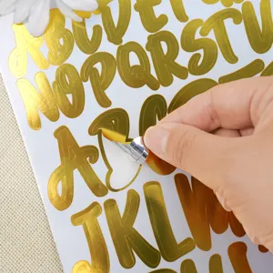 Close-up of a shiny gold foil alphabet sticker being peeled from its white backing sheet.