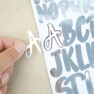 Close-up of a hand peeling a silver metallic alphabet sticker from the sheet