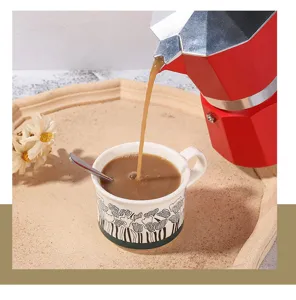 Classic red aluminum moka pot detail shot pouring fresh espresso into patterned ceramic coffee cup.