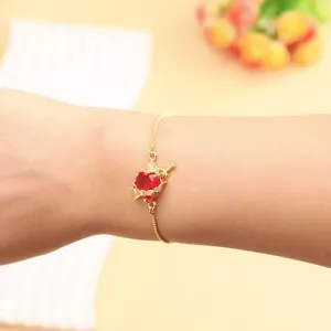 Detail shot of a gold-plated adjustable bracelet featuring a red heart and dagger pendant.