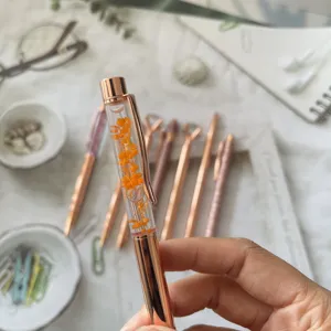 Rose gold dried flower ballpoint pen detail, transparent liquid barrel with vibrant orange floral elements.
